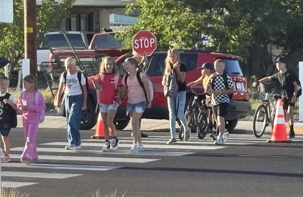 A group of children walk and ride bikes across a crosswalk after school. The kids wear backpacks, some chatting and smiling, while others push bicycles. A red SUV and stop sign are visible in the background.