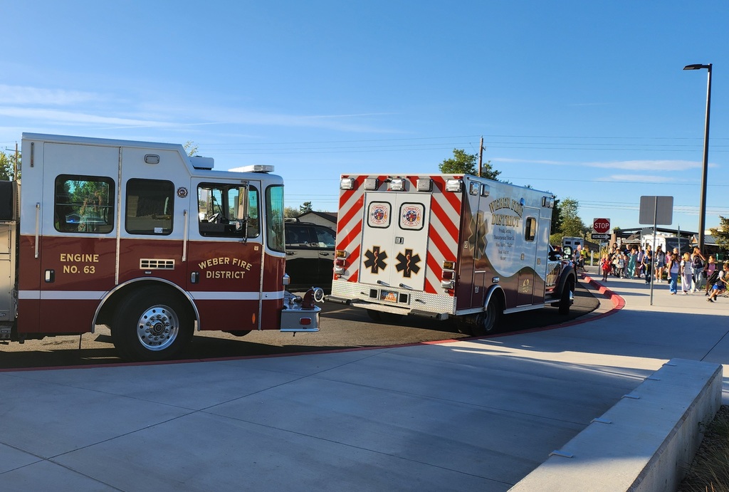 A Weber Fire District fire engine and ambulance are parked near a school curb on a clear sunny day. In the background, a crowd of children and parents are gathered on the sidewalk near a stop sign.