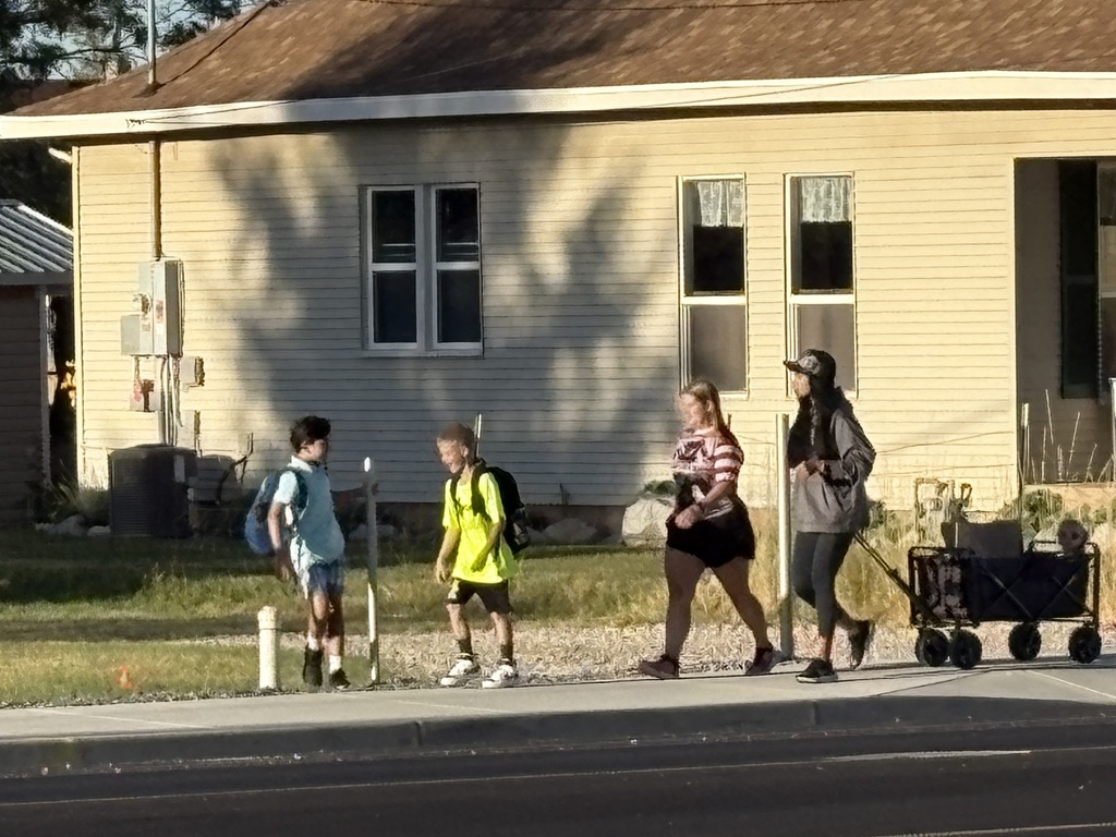 Several children walk along a sidewalk past a beige house after school. Two boys with backpacks talk near the grass, while two girls walk side by side. Another child pulls a wagon with a small child sitting inside.