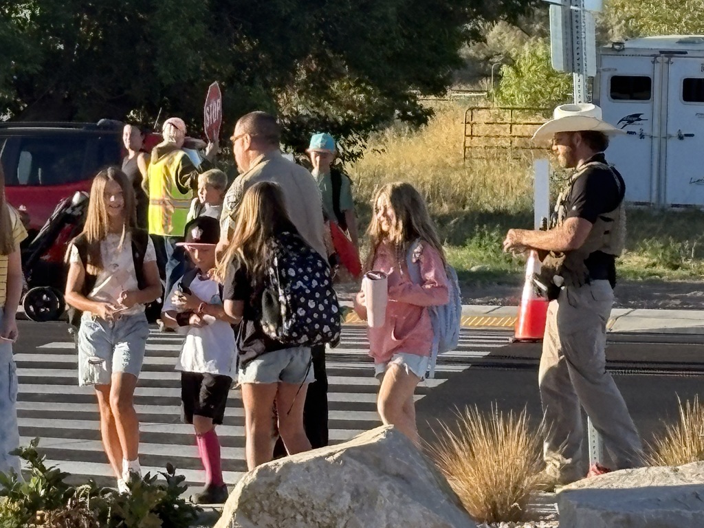A group of children with backpacks cross a street, smiling under the supervision of adults in safety vests and Weber County Sheriffs. The atmosphere is lively and safe.