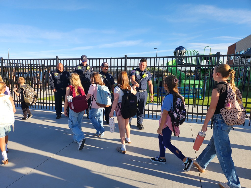 Children with backpacks walk past friendly firefighters in uniforms by a school fence. The scene is sunny and cheerful, conveying a sense of safety.
