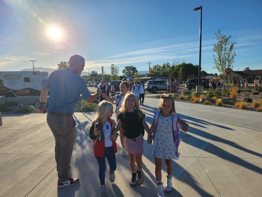 A group of smiling children walk on a sunlit path, accompanied by an adult. The clear blue sky and long shadows indicate early morning.