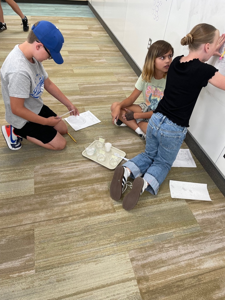 Three children work on a project, sitting and kneeling on a classroom floor. They have papers and glass jars, indicating a science experiment.