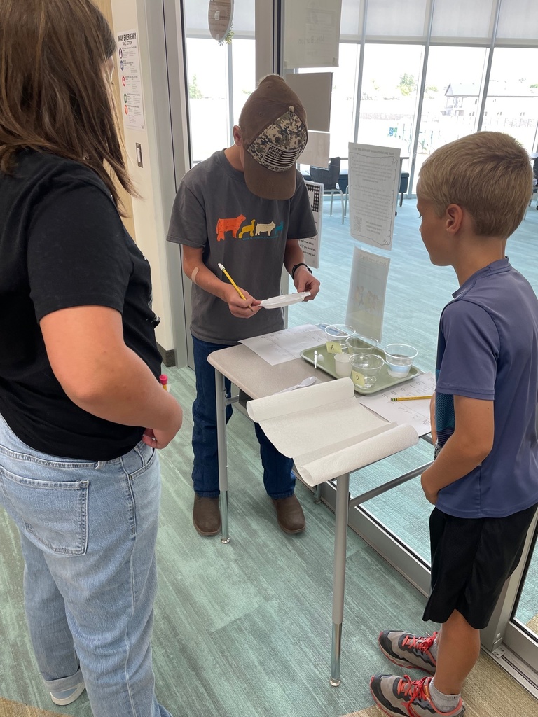 Three students engage in a science activity near a window. One writes notes while others observe. Materials include paper, bowls, and a pencil on the table.