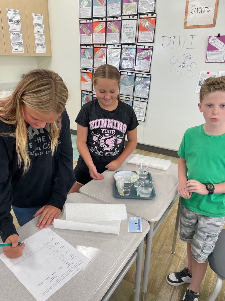 Three children in a classroom conduct a science experiment. One writes on a sheet, another observes with a smile, and the third looks forward. The setting is engaged and educational.
