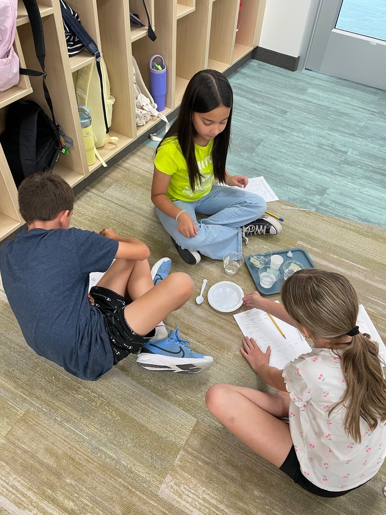 Three children sit on a classroom floor, engaged in a group activity with papers, plastic utensils, and cups, suggesting a collaborative task.