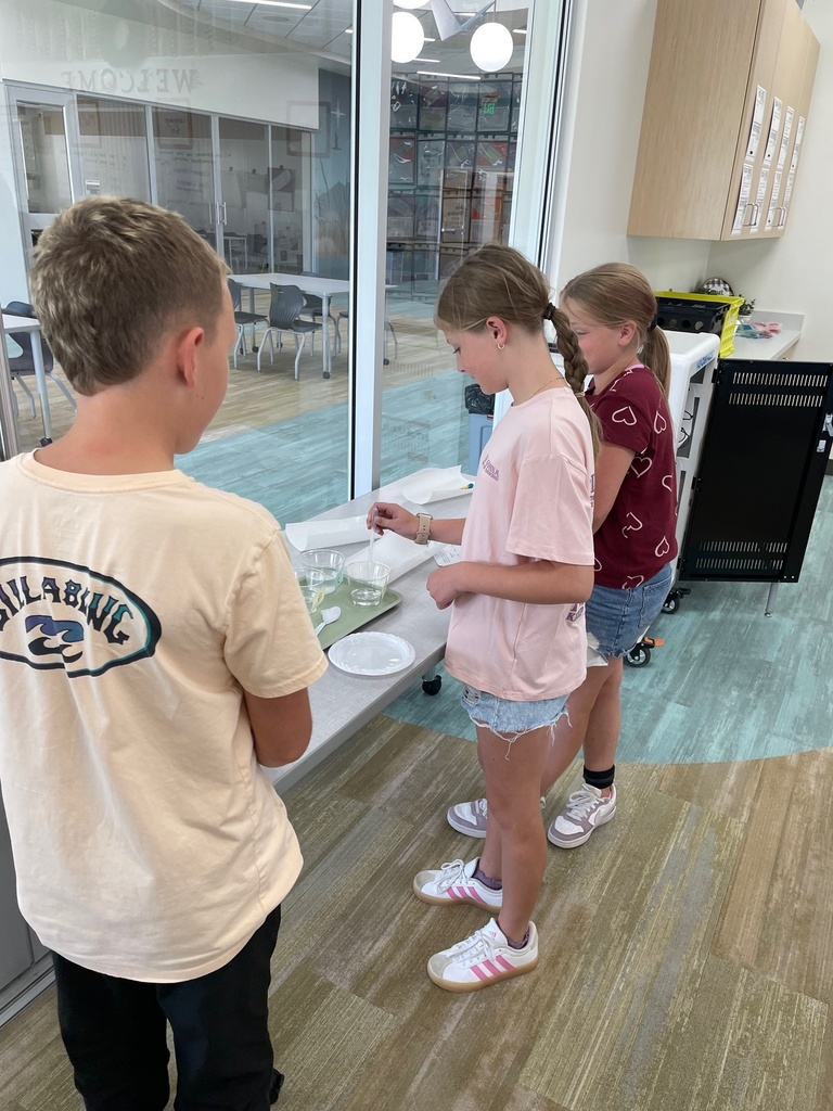 Three children stand at a table in a bright room, engaged in a science activity with liquids in bowls. They appear focused and intrigued.