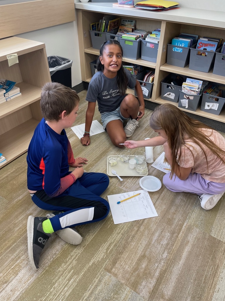 Three children sit on the classroom floor, engaged in a science experiment. They have a tray with cups in front. Shelves with books and bins are in the background.