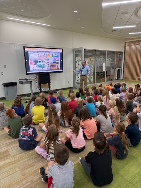 Students seated on the floor of a bright school commons face a large display while an adult presenter gestures toward an image-filled slide. The screen shows a colorful layout, and glass-walled classrooms line the back wall.