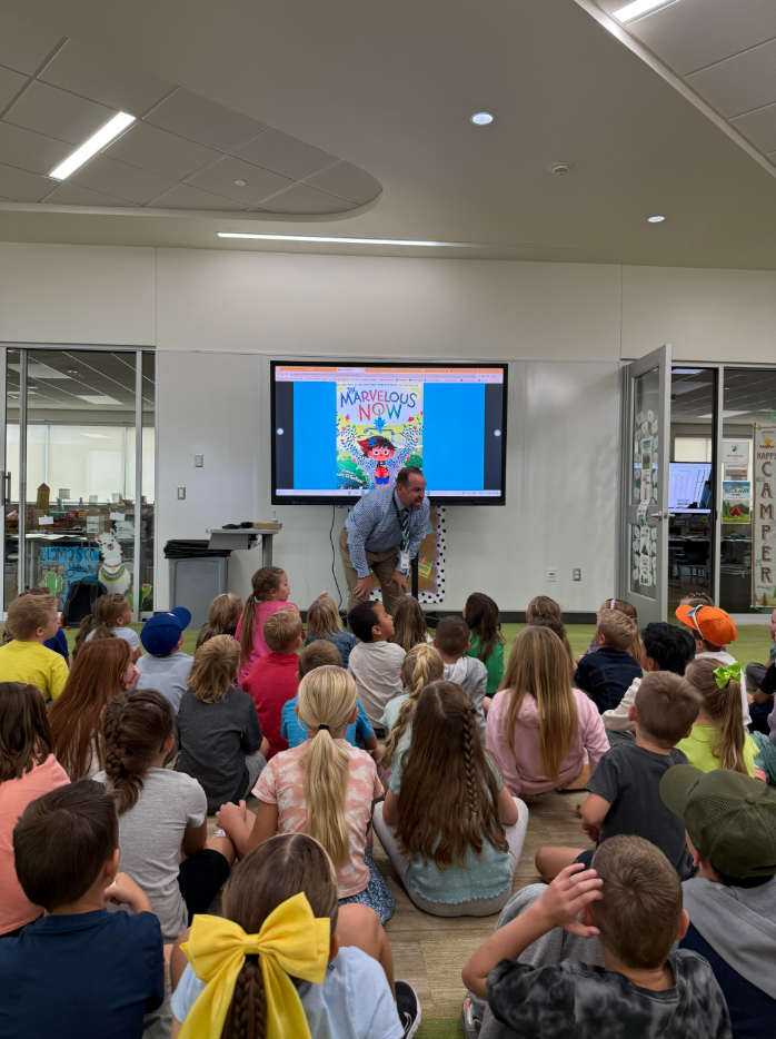 District Elementary Assistant Director crouches to speak to a large group of seated elementary students while a screen behind shows a colorful book cover titled “Marvelous Now.”