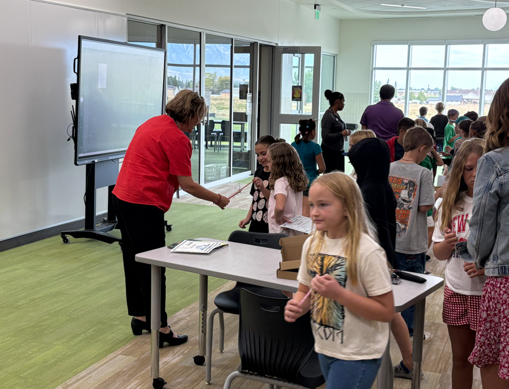 The Superintendent in a red shirt hands out items to elementary students lined up in a bright classroom with large windows and a digital display.