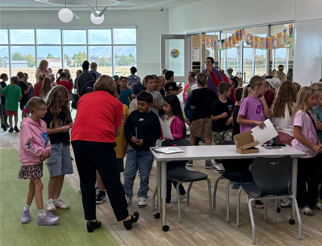 Dozens of elementary students gather and talk in a sunny classroom; the Superintendent  in a red shirt assists near a table with papers and boxes.