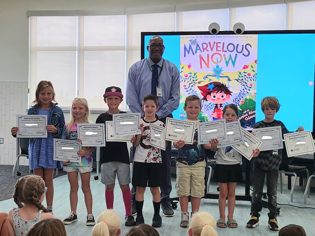 Seven elementary students stand in a row on a small stage area holding printed certificates, smiling for a photo. Assistant Superintendent, Clyde Moore, stands behind them, and a large screen displays the “The Marvelous Now” book cover as a backdrop. Students in the audience are visible in the foreground.