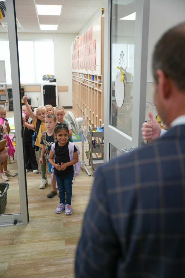 A group of young kindergarten students lines up at a classroom doorway, smiling toward an adult greeter in a suit who gives a thumbs‑up.