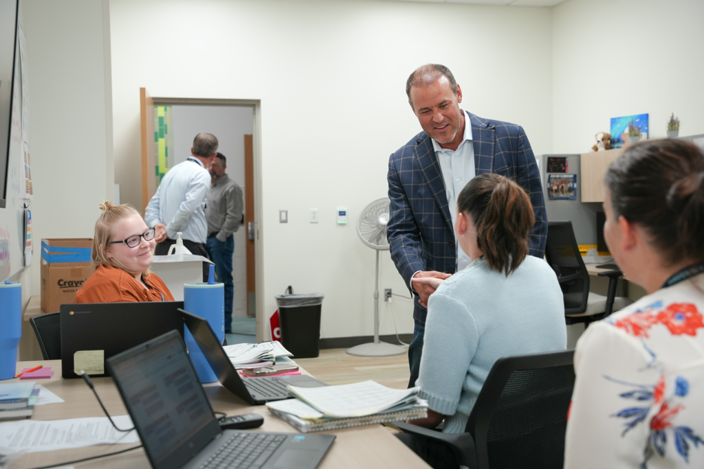 Speaker of the House Mike Schultz shakes hands with a staff member inside an office space at Haven Bay Elementary. Other staff members sit nearby at desks with laptops and notebooks, smiling and engaged. Two people are visible in conversation in the doorway in the background.