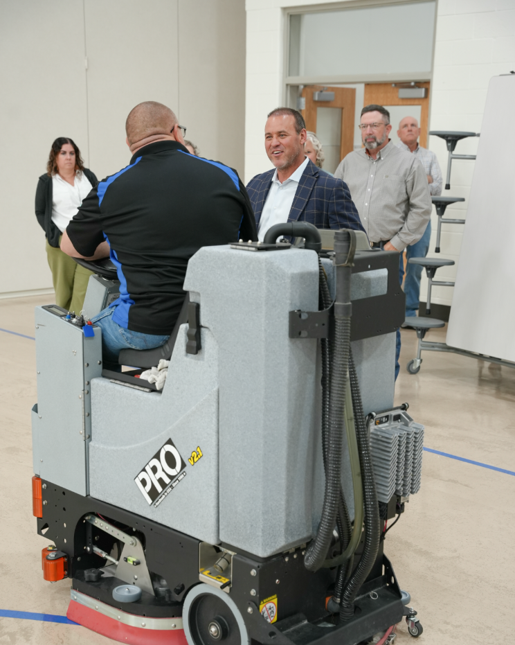 In a school cafeteria, a facilities staff member sits on a large ride‑on floor scrubber while visitors stand nearby and chat during a tour.