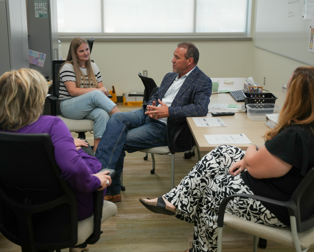Mike Schultz sits in a chair during a small group discussion with Haven Bay staff members in an office. Three staff members sit in a circle around him, listening attentively. Papers, folders, and a computer are on the table nearby.