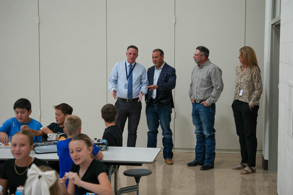 Mike Schultz stands in the lunchroom at Haven Bay Elementary alongside school and district leaders. Several students are seated at tables in the foreground, eating lunch, while Schultz and others talk and observe from the side.