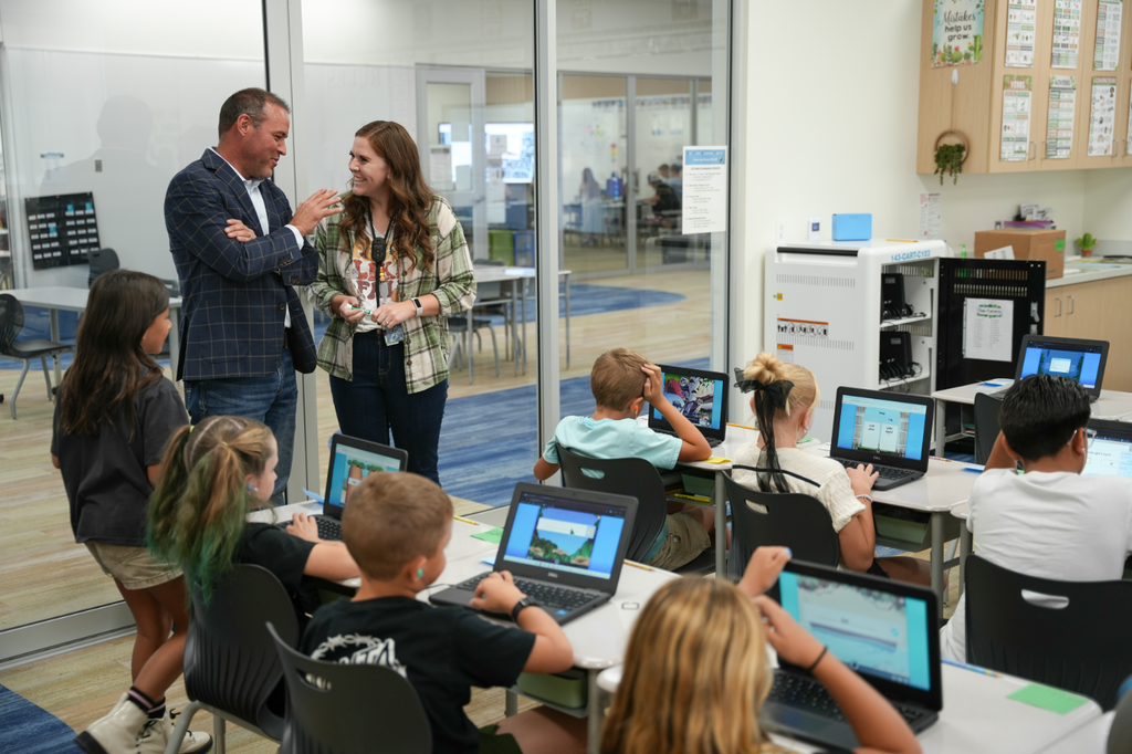 Elementary students work on laptops at tables while an adult visitor speaks with the teacher at the front of the classroom; glass walls reveal adjoining learning spaces.