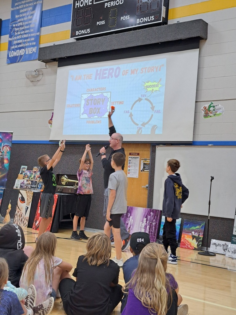 Author presenter on stage interacting with three elementary students who are raising their hands, in front of a projection screen showing a 'Story Box' diagram and 'Hero of My Story' title.