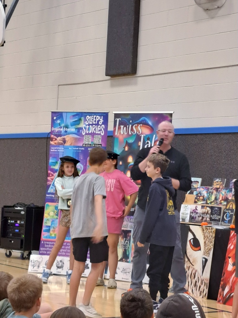 Author speaking into a microphone on stage, engaging with several elementary students who are standing near him, with colorful author banners in the background.