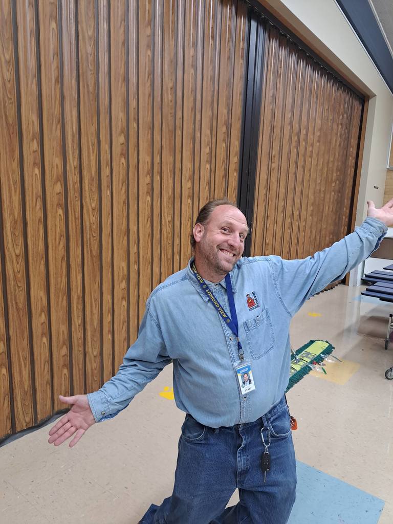 Smiling male school custodian kneeling down with arms outstretched in front of a folded-up divider wall.