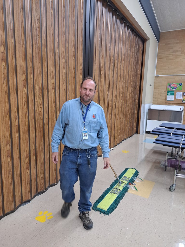 Male school custodian in a blue shirt and jeans with a large floor mop in the school cafeteria. 