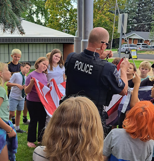 A male police officer in a dark blue uniform, seen from behind, demonstrates how to fold the American flag to a group of diverse elementary school-aged children gathered around a flagpole on a grassy area. The children are attentively watching the officer. 