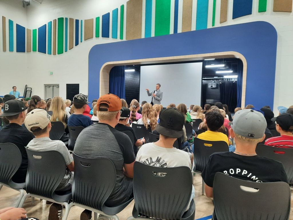 A group of 4th–6th grade students sit in an auditorium at Haven Bay Elementary listening to U.S. Representative Blake Moore, who is standing at the front speaking into a microphone. He is addressing the students about government while they sit in rows of gray chairs, many wearing hats. The stage behind him has a large projection screen, and colorful sound panels decorate the walls.