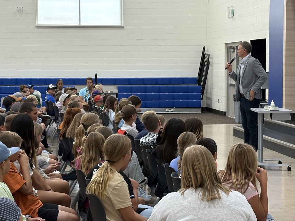 Side view of a school assembly showing a speaker with a microphone engaging a full audience of students seated in rows.