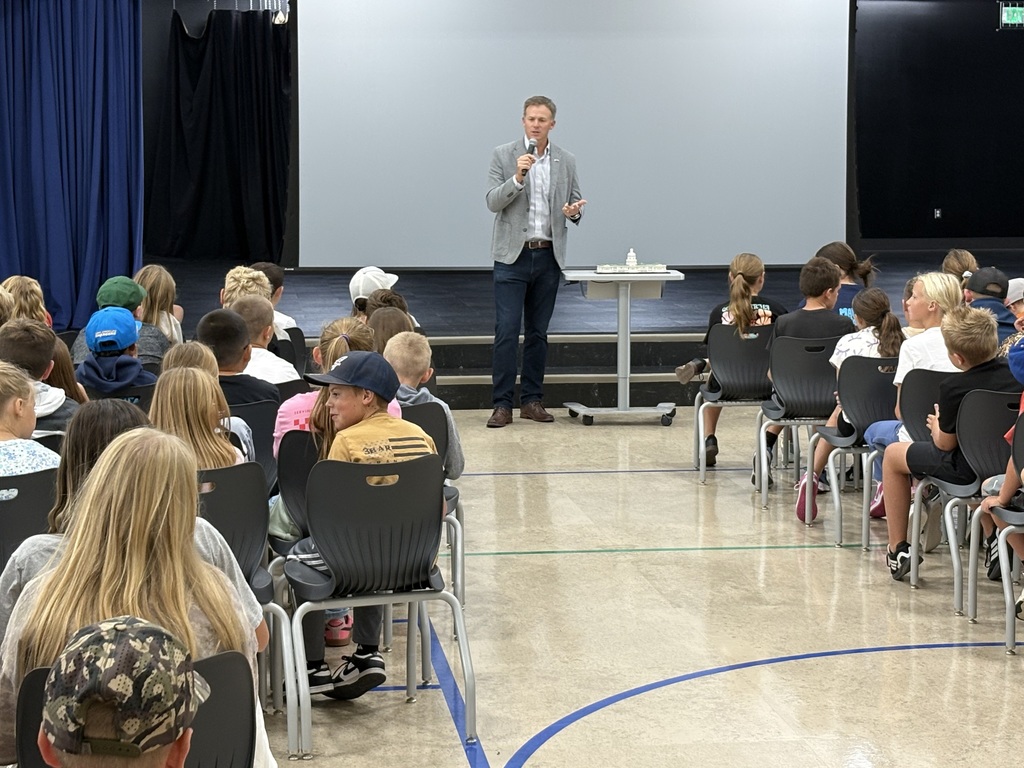 Rep. Blake Moore stands at the front of a multipurpose room with a microphone, speaking to rows of seated students facing the stage.