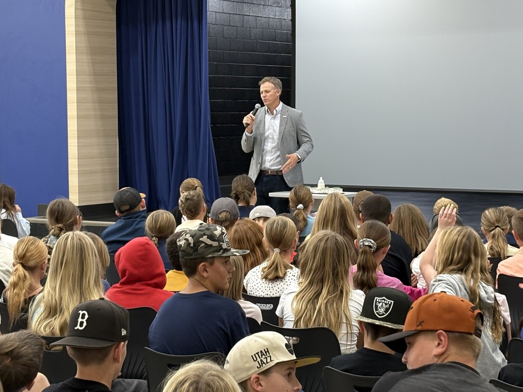 Closer view of the stage area where a speaker talks into a microphone while students seated up front listen and raise hands.