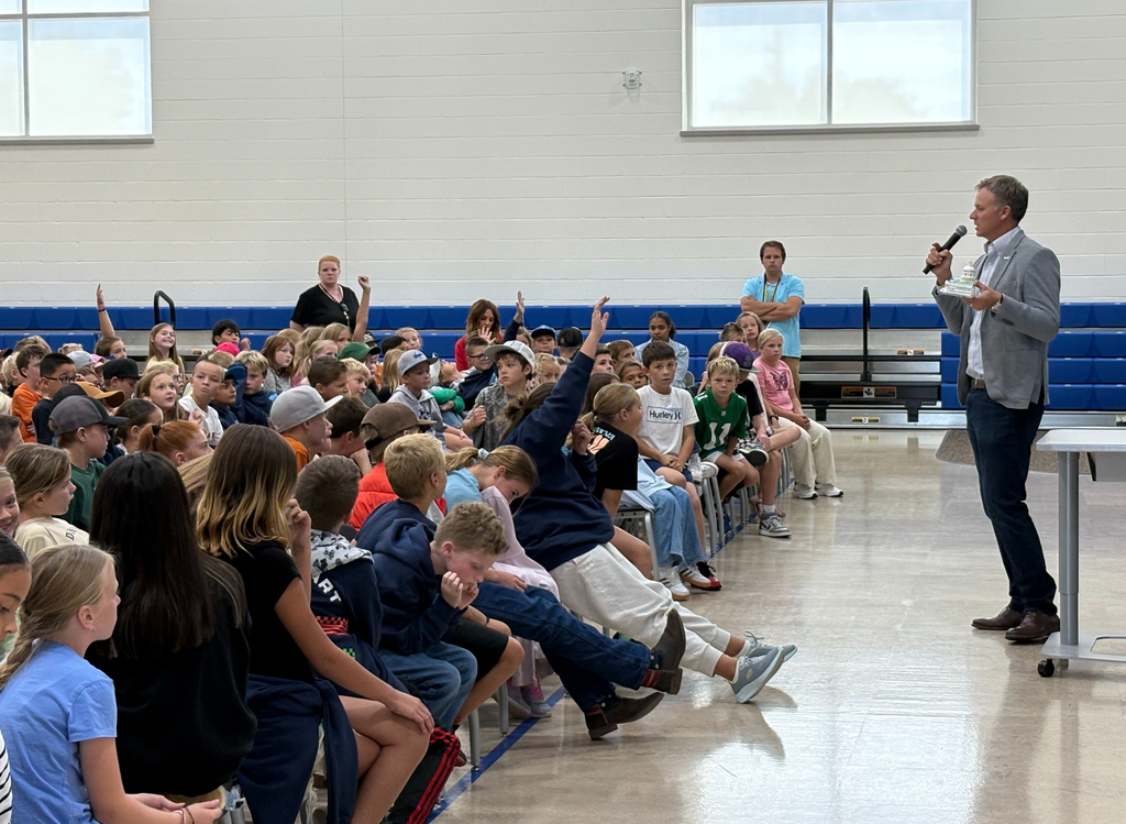 Rep. Blake Moore  with a microphone addresses a large group of elementary students in a gym; several students raise their hands to ask questions.