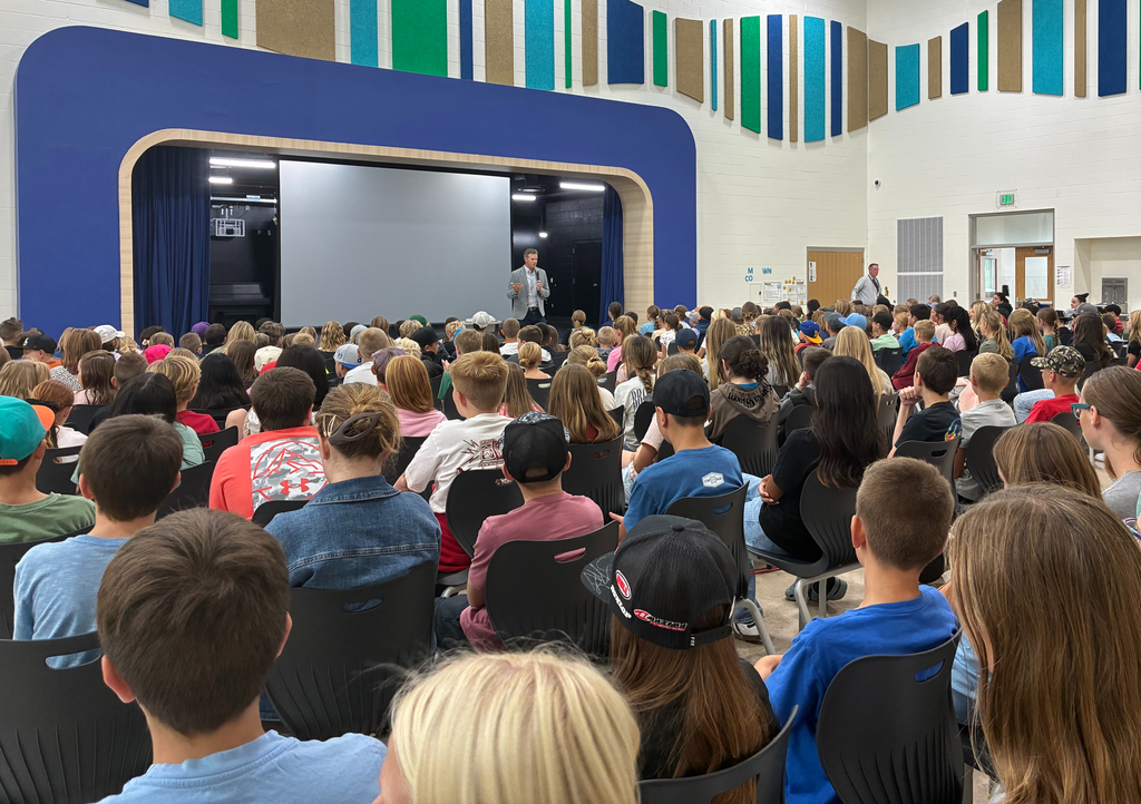 Wide view of a school assembly in a colorful auditorium as a speaker presents on stage to a packed audience of students.