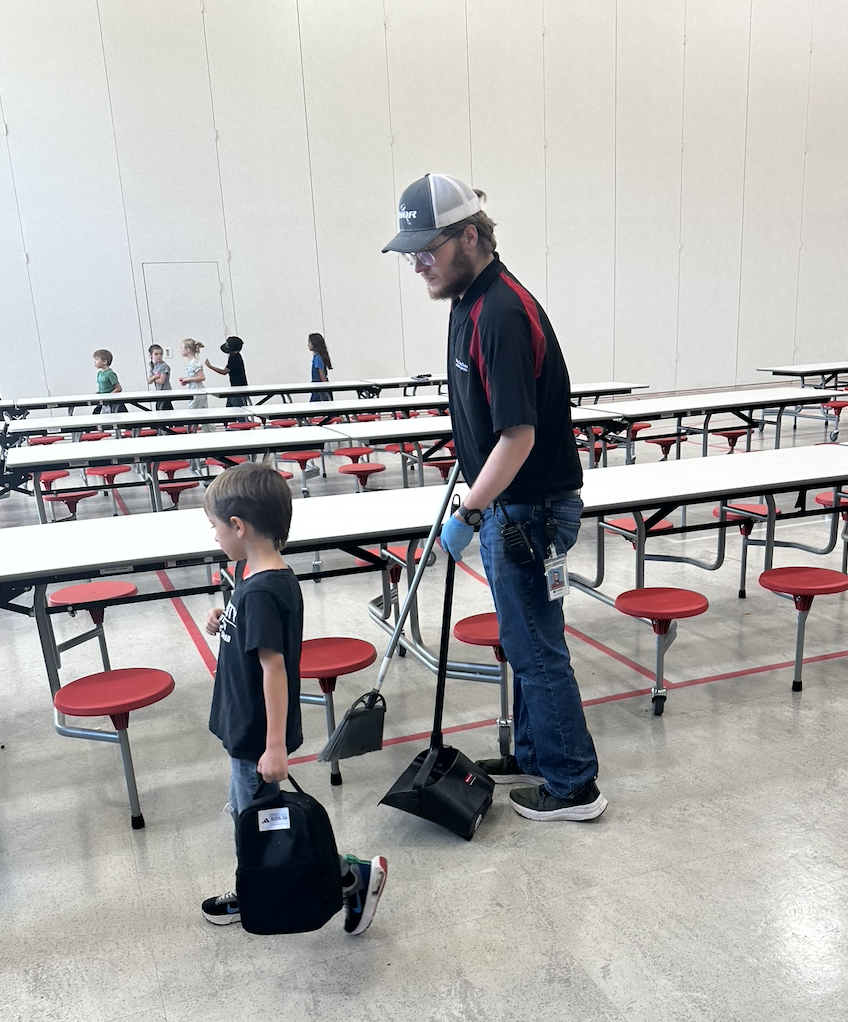 custodian sweeping trash up off the floor in the lunchroom, student in the foreground