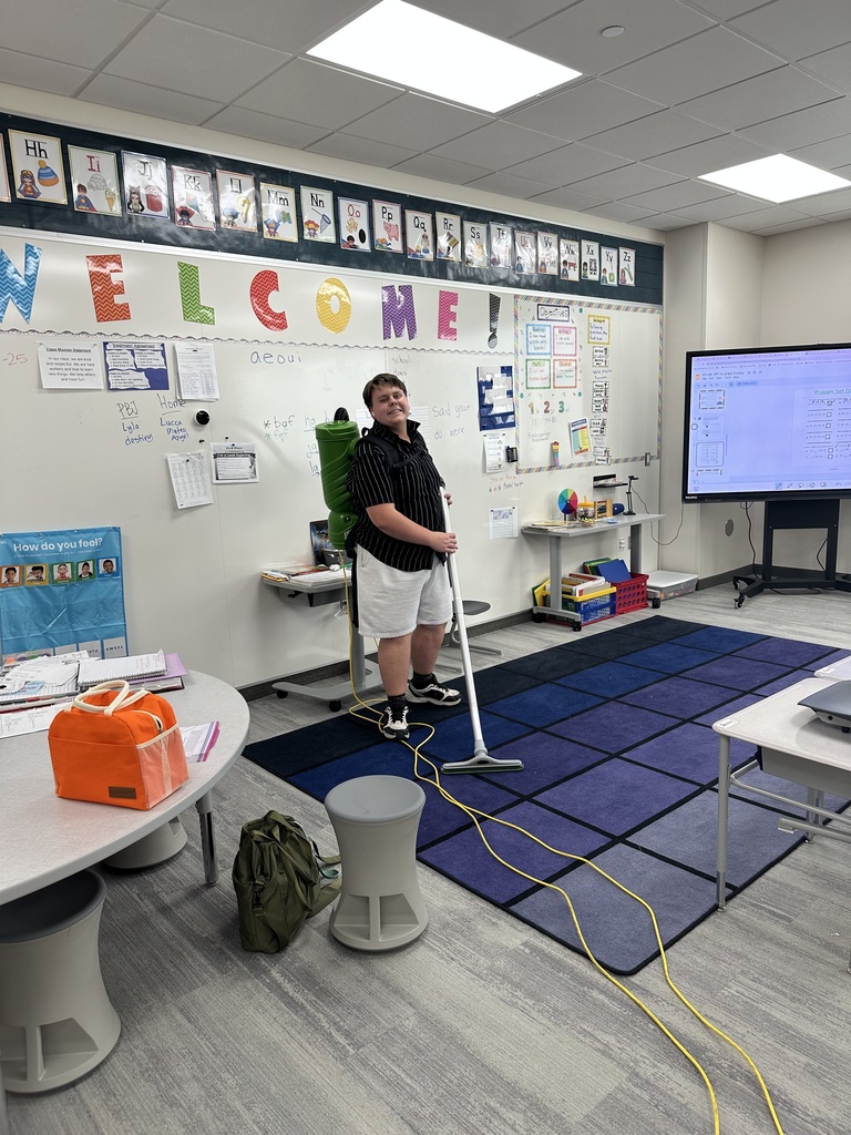 teenage custodian vacuuming the floor in a 1st grade classroom- he is smiling 