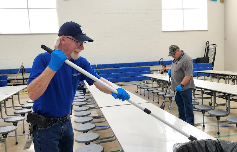 Two custodians wearing blue gloves clean a large, empty school cafeteria. The man in the foreground, wearing a blue shirt and baseball cap, smiles while using a long-handled tool to wipe down a white folding table. Another custodian in a gray shirt works on a table in the background. The room features high ceilings, long folding tables with attached gray stools, and blue padding along the wall.