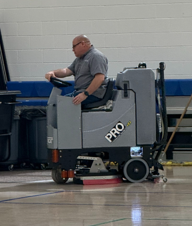 A custodian in a gray polo shirt is driving a large, gray, ride-on floor scrubber across the gymnasium or cafeteria floor. The machine has 'PRO v2.1' printed on the side. The man is focused on operating the equipment, which is cleaning the polished floor surface next to a wall with blue padding.