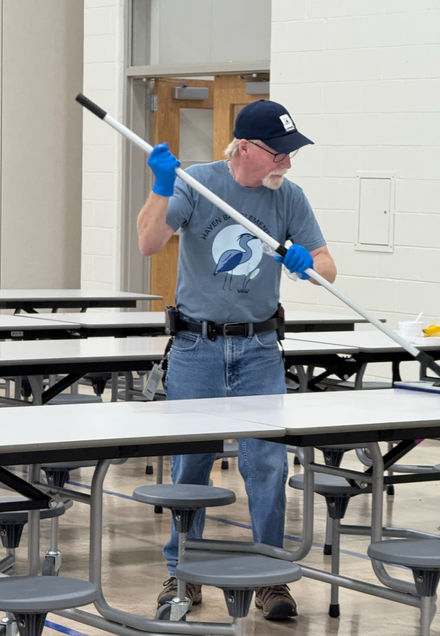 A custodian in a gray polo shirt is driving a large, gray, ride-on floor scrubber across the gymnasium or cafeteria floor. The machine has 'PRO v2.1' printed on the side. The man is focused on operating the equipment, which is cleaning the polished floor surface next to a wall with blue padding.