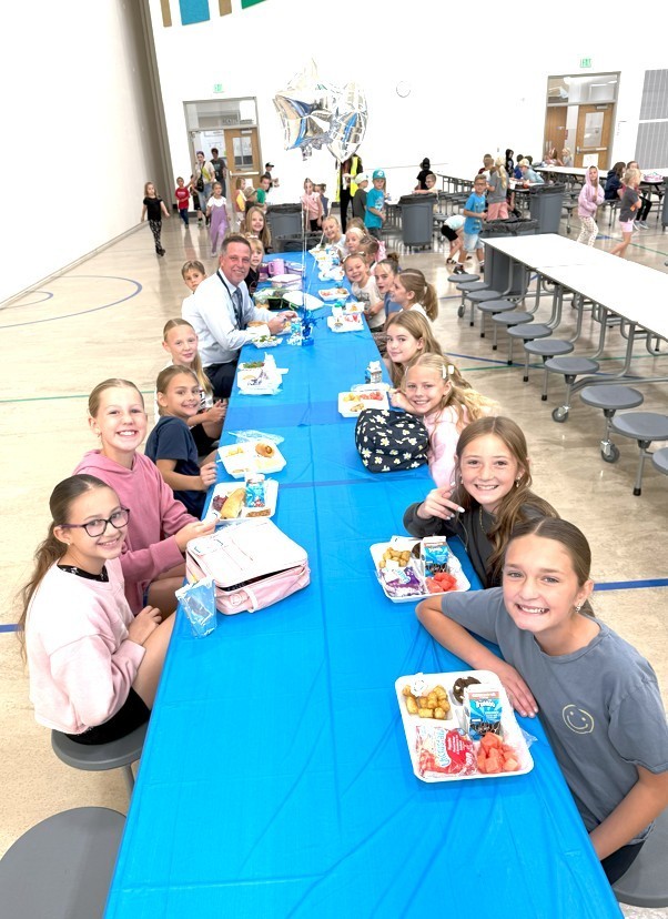 A wide-angle view of a group of smiling elementary school students and a male teacher seated at a long, light-blue covered table in a school cafeteria or gymnasium for a lunch celebration.