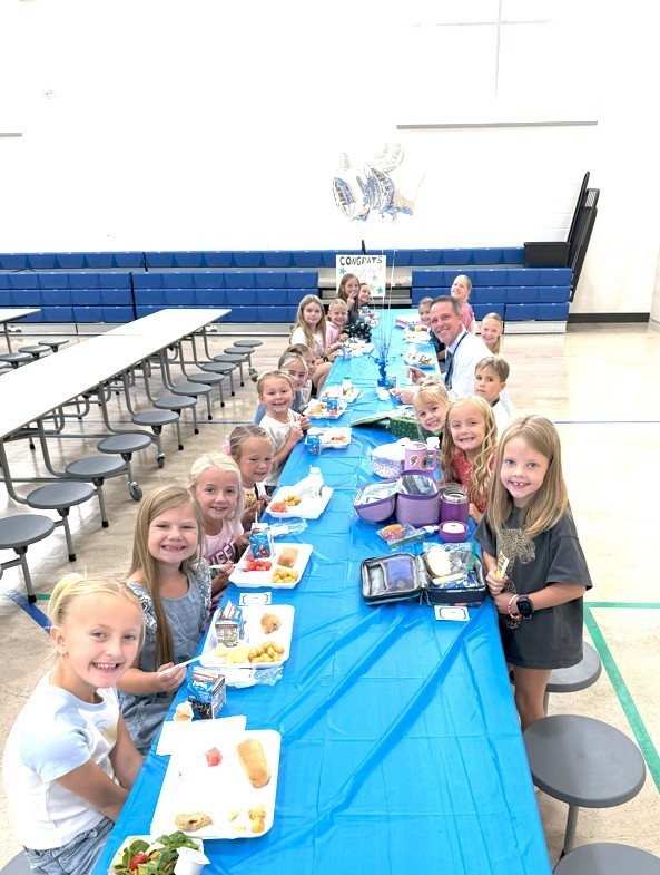 A long, light-blue covered table in a school gymnasium, with many smiling elementary school students and a male teacher seated for a lunch celebration. A balloon that says CONGRATS is visible in the background.