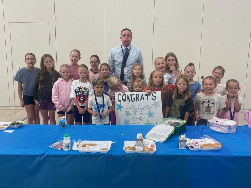 A group photo of about 15 elementary school students, mostly girls, and a male teacher standing behind a table. The children are making silly faces and holding a sign that says CONGRATS Students of the Month!. School lunch trays are on the blue-covered table.