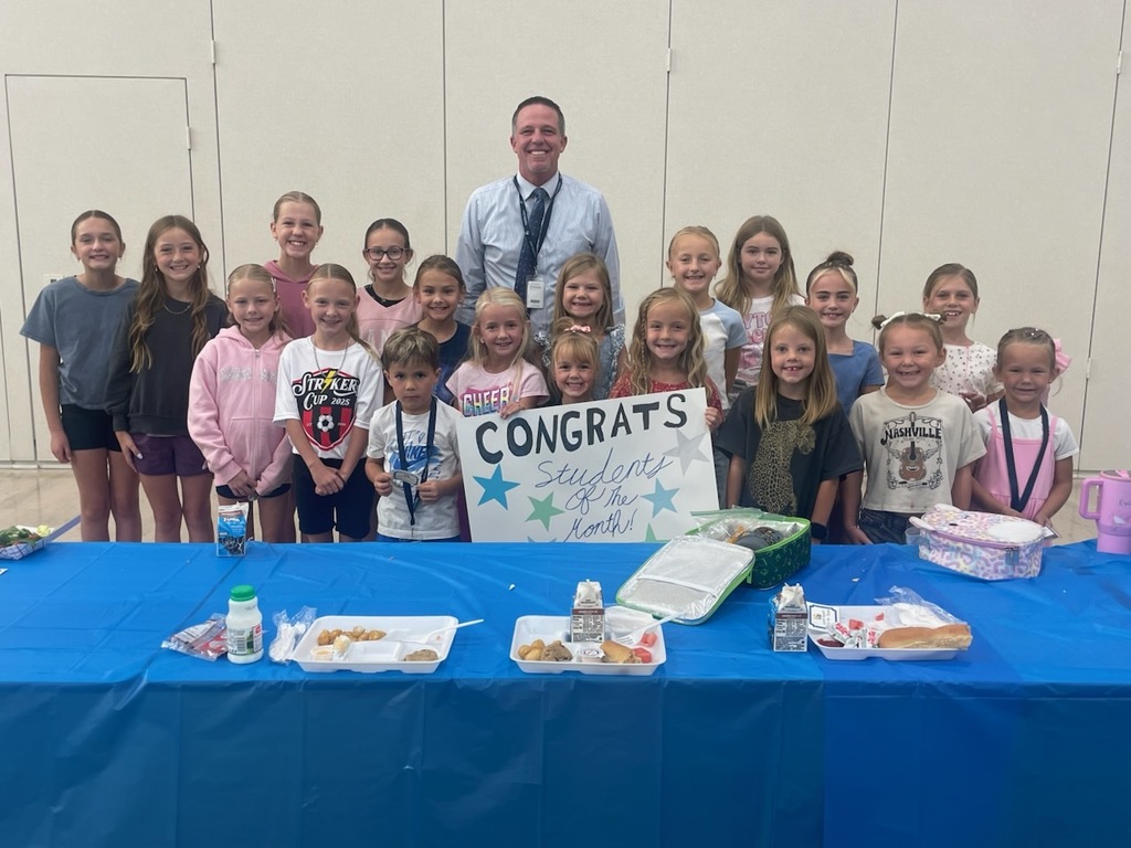 A group photo of about 15 smiling elementary school girls and a few boys standing behind a table with a male teacher, holding a sign that says CONGRATS Students of the Month!. School lunch trays and lunch boxes are on the blue-covered table in front of them