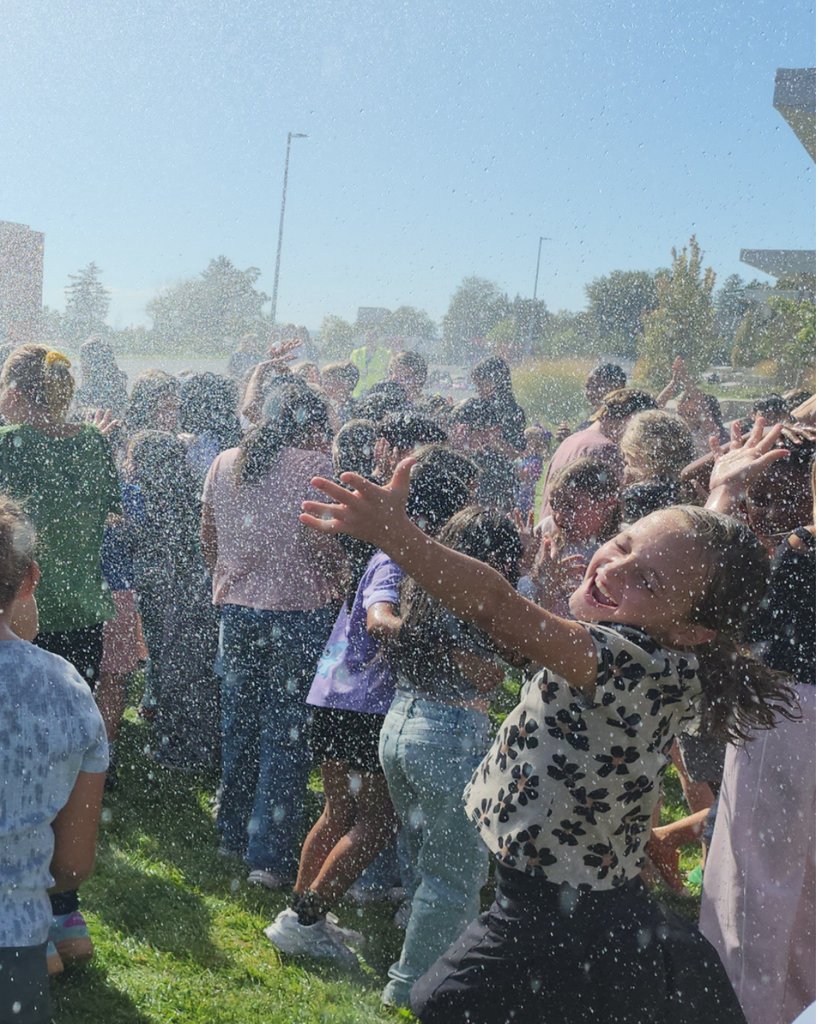 A close-up, sunlit photo of a joyful young girl with her arms outstretched and eyes closed in delight as she is sprayed with water. In the background, a crowd of other children are similarly enjoying being sprayed.