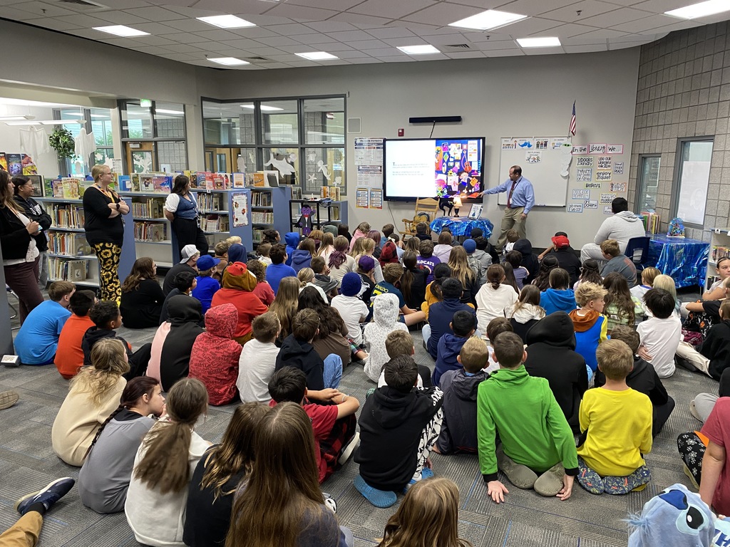 Students sitting and listening to a book read by a special visitor from the superintendents office.