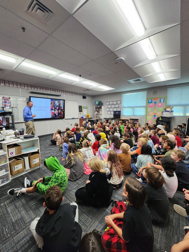 Students sitting and listening to a book read by a special visitor from the superintendents office.