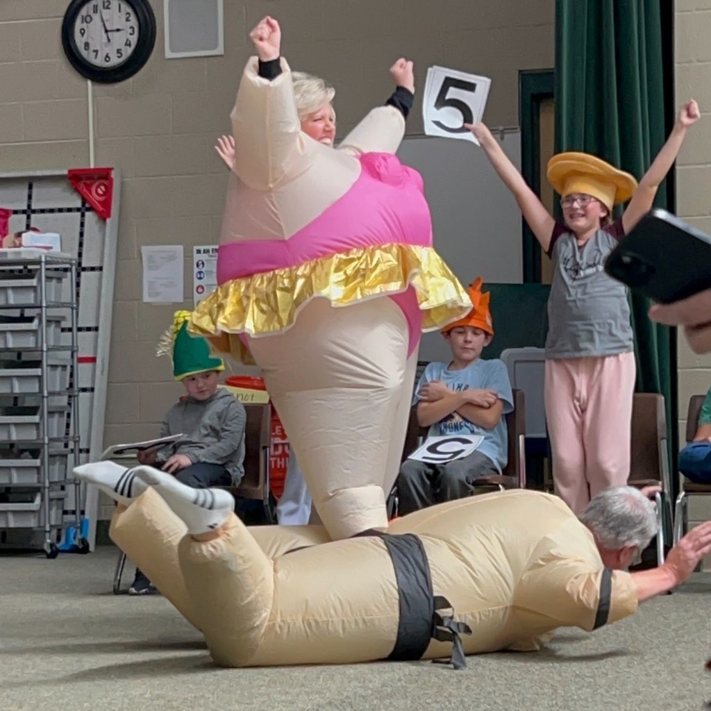 Mrs. Mower dressed in a Sumo costume stomping on Mr. Mower who is laying face down on the floor in a Sumo costume.