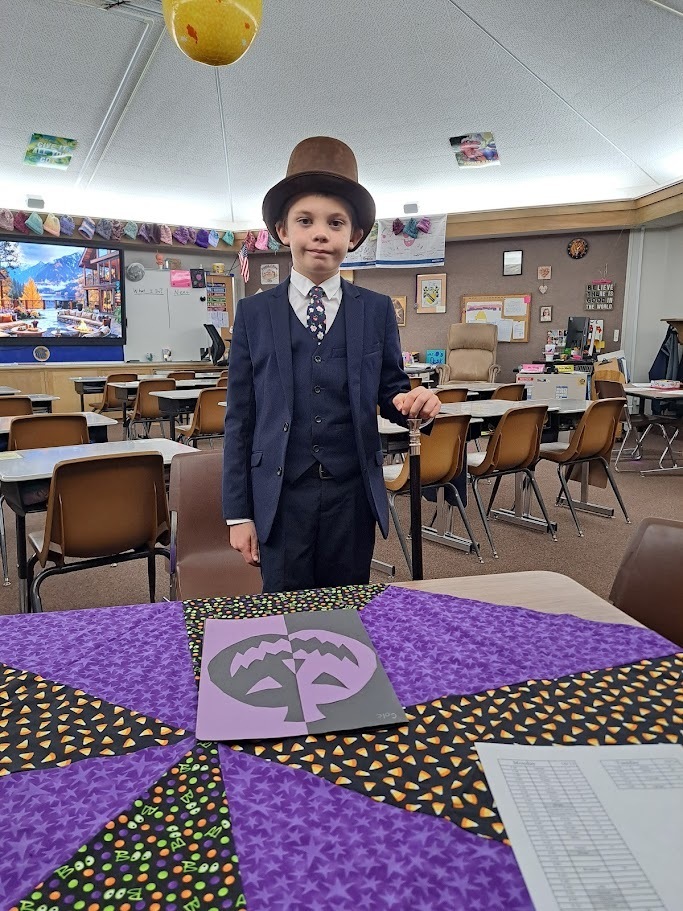 A young elementary school student stands proudly in his classroom wearing a full navy blue suit, vest, patterned tie, and a brown top hat. He is standing behind a table with a purple and black Halloween-themed quilt and a jack-o'-lantern art project on it.