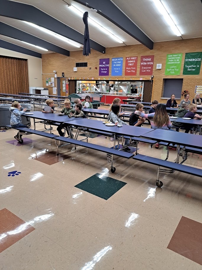 Elementary students in the Science Club sit at long blue tables in the school cafeteria, engaged in a hands-on learning activity about chemical reactions. In the background, colorful posters with leadership principles hang on a brick wall.