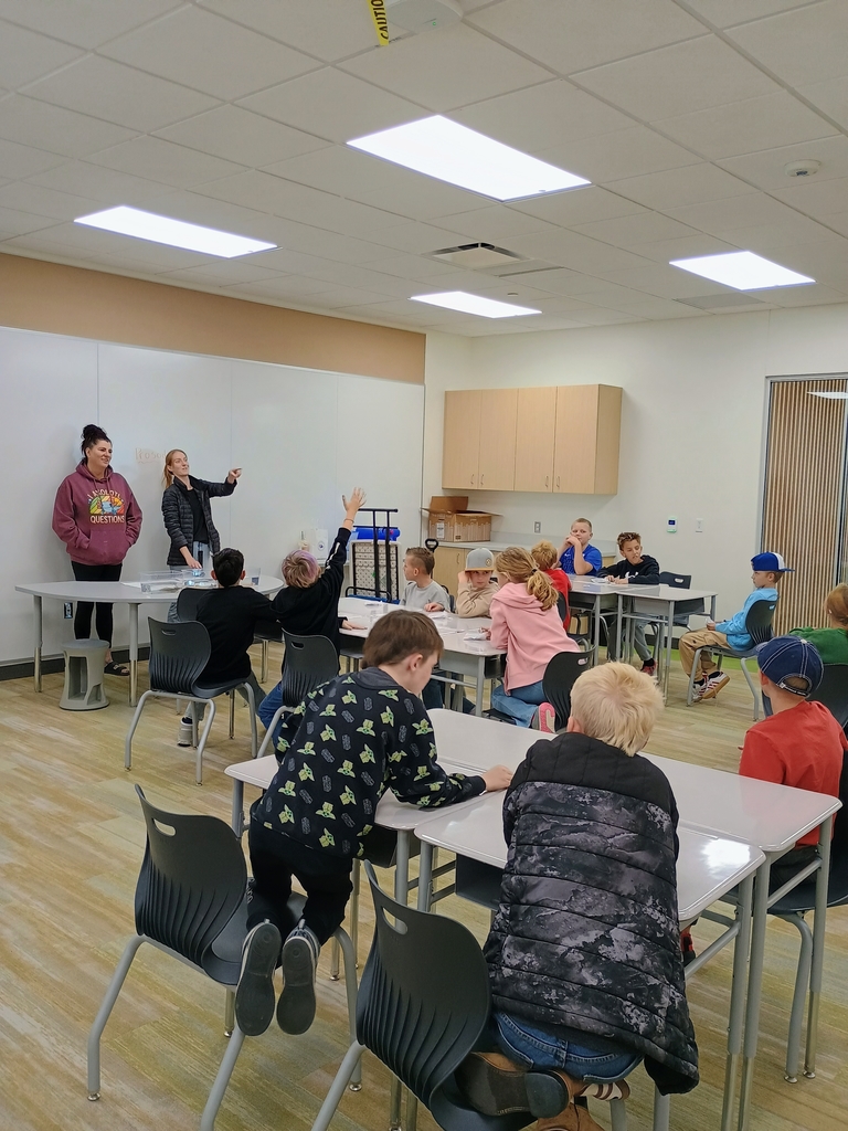 A classroom scene with elementary students seated at tables. Two adult women stand facing the class near a white wall. A boy seated in the middle raises his hand to ask a question.